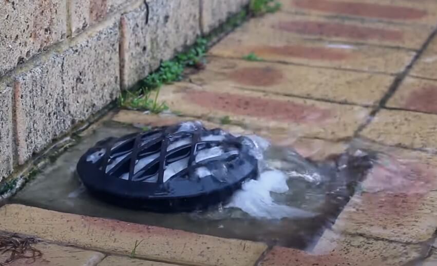 A dark gray drain cover sits in a shallow trench filled with water