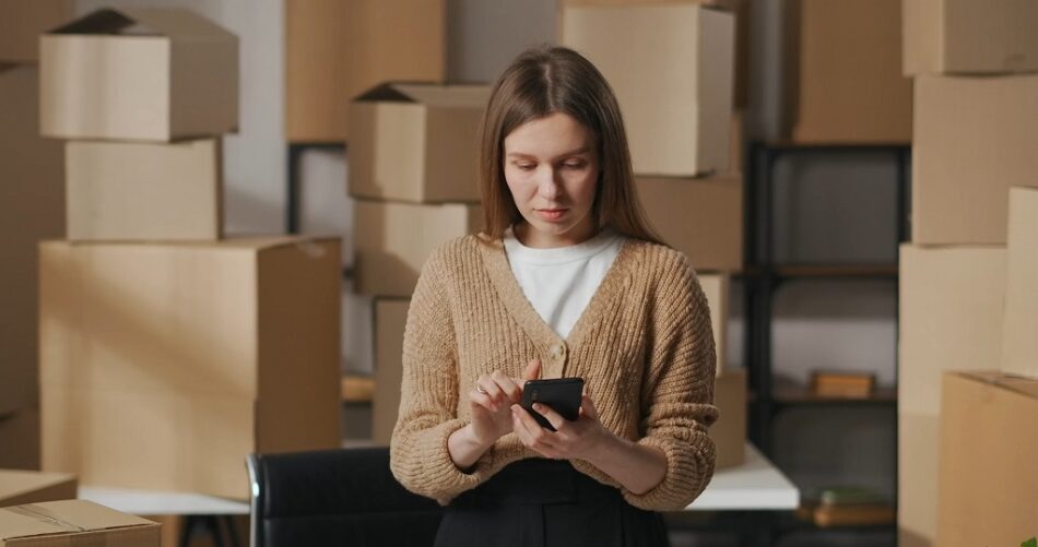 Ecommerce business owner managing orders on a smartphone while surrounded by packed shipping boxes in a small warehouse workspace
